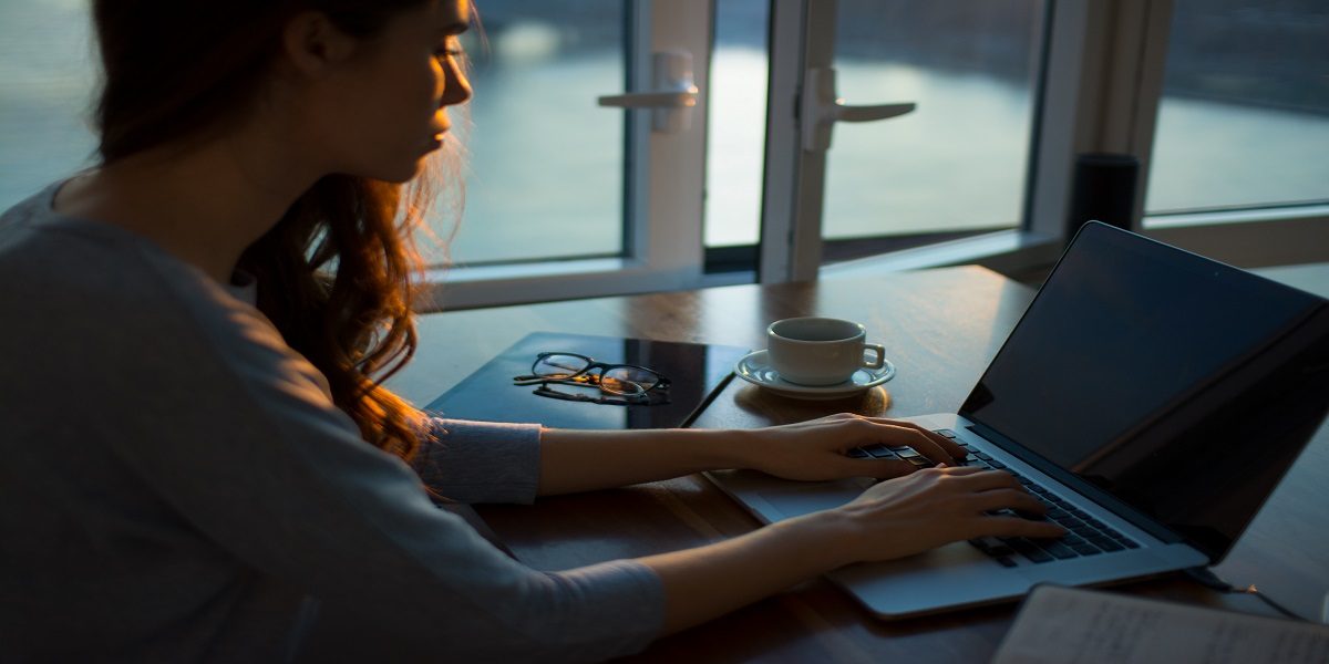 A woman sits by a window while typing on her laptop