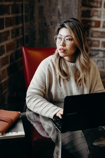 An East Asian woman in a cream sweater and glasses looks sideways while sitting down at a black laptop computer. Mental Health.
