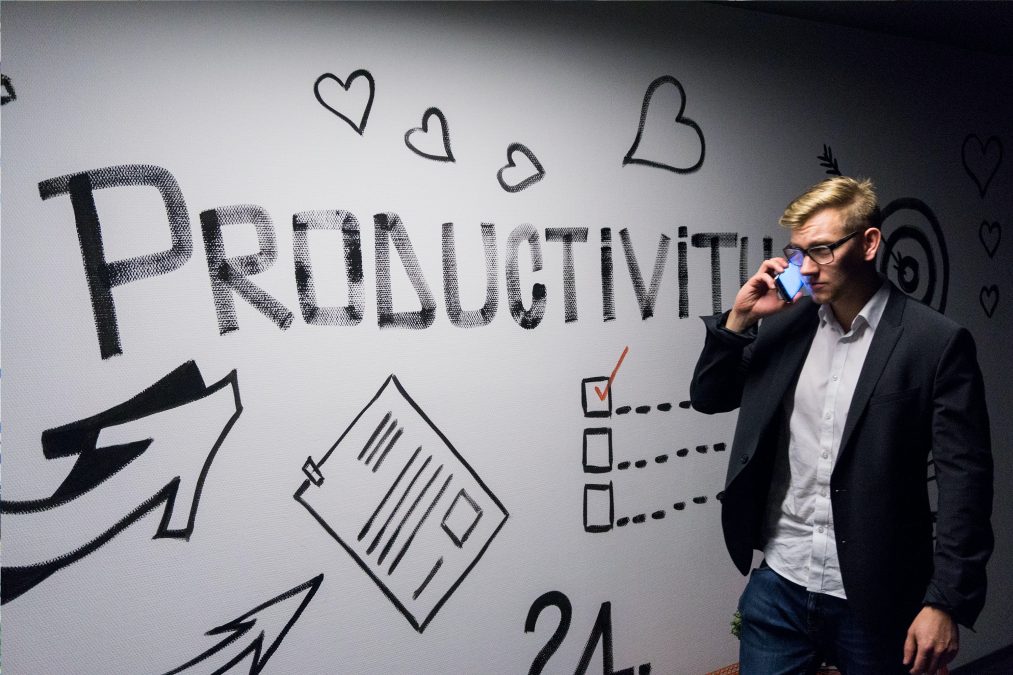 A white man with glasses, a white button-up shirt, a black blazer, and jeans is walking with a phone to his ear. To his right is a white board with big black block letters that say PRODUCTIVITY For the blog post How to Better Respond to People When You're Busy" by Deborah Beckwin