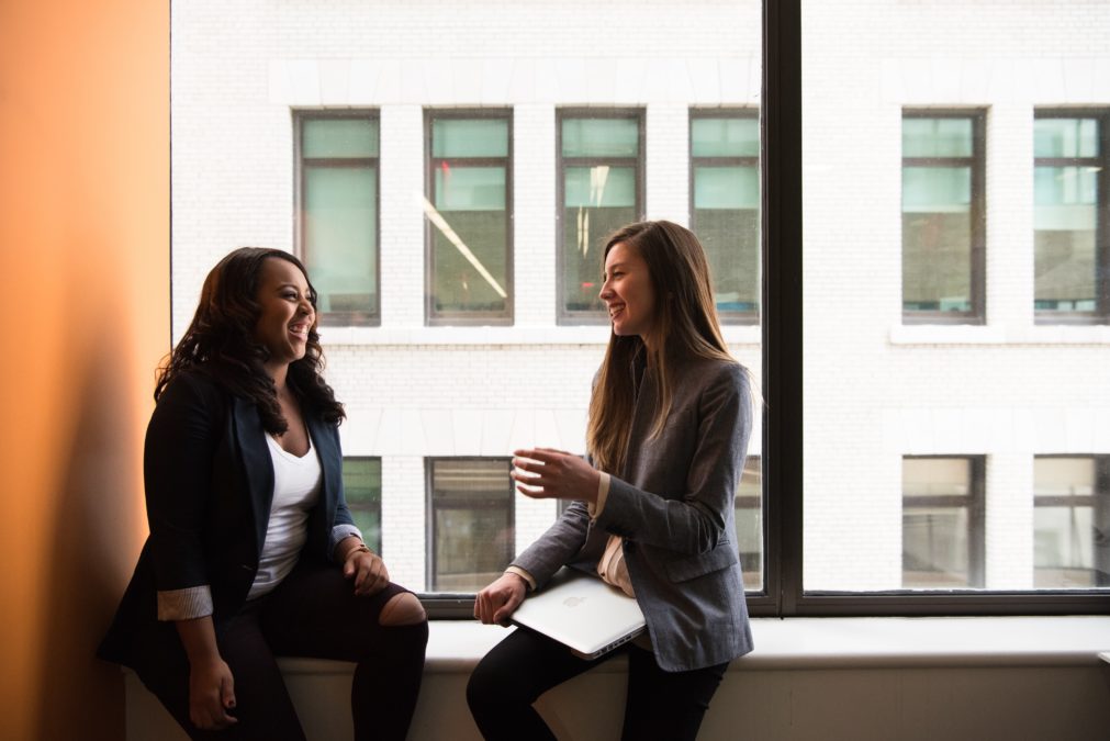 Two business women of color sit on a window sill smiling and talking