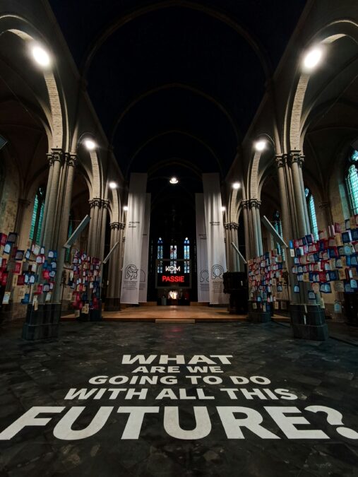 Interior of a church exhibition with lettering in white capital letters "what are we going to do with all this future?